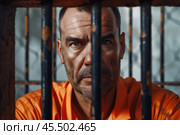 A serious prisoner in an aged orange uniform gazes directly at the camera from behind metal bars in a detention center, reflecting the harsh realities of incarceration. Стоковое AI-изображение, AI-художник Sergey Grishin / Фотобанк Лори