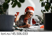 Woman in Santa hat enjoys cozy Christmas at home, relaxing with smartphone during holiday season. Surrounded by festive warmth, she uses smartphone to connect and celebrate Christmas moments. Стоковое фото, фотограф Енваев Андрей / Фотобанк Лори