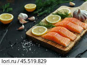 Salmon Fillet. Fresh salmon fillet with parsley and lemon slices. Sliced raw pink salmon fish steak with cooking ingredients, herbs on a wooden cutting board on a dark black background, top view. Стоковое AI-изображение, AI-художник Марина Демидюк / Фотобанк Лори