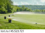 Sprinkler system irrigating green grass field on a sunny day. Стоковое AI-изображение, AI-художник Юрий Поздников / Фотобанк Лори