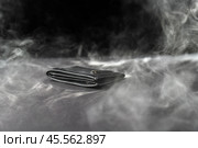 Black leather wallet resting on a dark backdrop, surrounded by swirling smoke. Money appears to be burning, symbolizing fleeting wealth and the transient nature of material possessions. Стоковое фото, фотограф Татьяна Степанищева / Фотобанк Лори