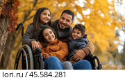 A happy family of four against the backdrop of an autumn landscape. The central element is the father, who is smiling and hugging his children. The eldest daughter is sitting in a wheelchair, her face glowing with joy. Around her, yellow leaves of trees create an atmosphere of warmth and coziness. The father and children are dressed in warm autumn clothes: sweaters and jackets. The picture conveys a sense of closeness, love and family happiness, and also demonstrates inclusivity and equality. Стоковое AI-изображение, AI-художник Анна Кривицкая / Фотобанк Лори
