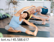 Old woman practicing seated side bend pose of yoga in training room during fitness classes. Стоковое фото, фотограф Яков Филимонов / Фотобанк Лори