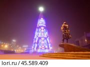 Красивая новогодняя ель в Якутске. Ночной городской пейзаж. На переднем плане памятник писателю Платону Ойунскому. Вдали на здании название Якутска на якутском языке — «Дьокуускай». Площадь Орджоникидзе, Город Якутск, Республика Саха (Якутия), Россия. Большая искусственная ёлка, украшенная традиционными якутскими орнаментами. Праздничное новогоднее настроение. Холодная погода и морозный туман. (2024 год). Редакционное фото, фотограф Андрей Степанов / Фотобанк Лори