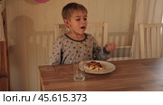 Cheerful child is enjoying a delicious breakfast in a cozy dining room. Young boy sits happily at a rustic wooden table, enjoying a scrumptious and delicious meal in a warm, inviting space. Стоковое видео, видеограф Марина Демидюк / Фотобанк Лори