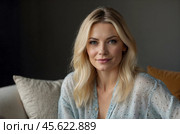 Head shot portrait pretty blonde woman looking at camera, sits alone on sofa photoshooting, enjoy pleasant pastime weekend at home at own or rented apartment. Beautiful mature female profile picture. Стоковое AI-изображение, AI-художник Александр Поташев / Фотобанк Лори