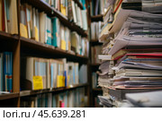 Shelves are lined with neatly organized folders and papers, while stacks of documents rise prominently in the center of the library, showcasing the busy atmosphere. Стоковое AI-изображение, AI-художник Sergey Grishin / Фотобанк Лори