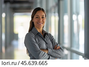 A Hispanic female facility manager stands with crossed arms, exuding confidence and approachability. She smiles warmly, set against a backdrop of a bright, modern corridor. Стоковое AI-изображение, AI-художник Sergey Grishin / Фотобанк Лори