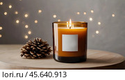 A mock-up of a cozy burning candle made of amber glass with an empty white label in a festive, New Year's home interior. Стоковое AI-изображение, AI-художник Photojuli / Фотобанк Лори