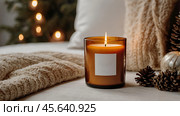 A mock-up of a cozy burning candle made of amber glass with an empty white label in a festive, New Year's home interior. Стоковое AI-изображение, AI-художник Photojuli / Фотобанк Лори