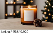 A mock-up of a cozy burning candle made of amber glass with an empty white label in a festive, New Year's home interior. Стоковое AI-изображение, AI-художник Photojuli / Фотобанк Лори