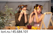 Two female friends sharing moment of relaxation and enjoying beauty routine at home, sitting by tabletop vanity mirror and curling hair with colorful rollers. Стоковое видео, видеограф Яков Филимонов / Фотобанк Лори