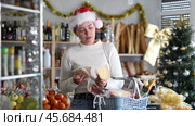 Thoughtful female customer in funny Santa hat standing in small festively decorated grocery store, examining packages of cheese, choosing goodies for Christmas celebration. Стоковое видео, видеограф Яков Филимонов / Фотобанк Лори