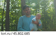 A bearded father in a blue jersey and black baseball cap holds a baby in a blue onesie against a forest background. A man smiles while playing with his son in the park holding him in his arms. Стоковое видео, видеограф Воловодов Владимир / Фотобанк Лори