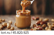 Spoon and glass jar with creamy peanut butter on kitchen table, closeup. Spoonful of peanut butter. Creamy Peanut butter and peanuts on the dark brown wooden table rustic background. Стоковое AI-видео, аниматор Марина Демидюк / Фотобанк Лори