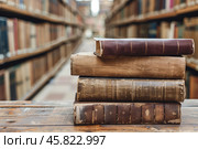 A stack of antique leather-bound books rests on a wooden table set against the blurred background of library bookshelves. Shallow depth of field. Стоковое AI-изображение, AI-художник Михаил Строгалев / Фотобанк Лори