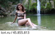 Gorgeous woman in mesh black bikini sits on rock on shore of tropical waterfall. Стоковое видео, видеограф Яков Филимонов / Фотобанк Лори