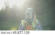 Woman wearing a safety vest and a hard hat is holding a device that looks like a camera. She is standing in a field with trees in the background. Стоковое видео, видеограф Скрипачев Антон / Фотобанк Лори