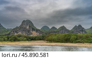 A picturesque mountain range along the river bank. The slopes are covered with green vegetation. Peaks against the sky and clouds. Trees and grass grow on the sandy shore. China. Li River. LiJiang. Стоковое фото, фотограф Вера Ивановна Кубарева / Фотобанк Лори