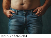 A male figure captured from the chest to the hips. The man is wearing classic blue jeans that fit his figure slightly. The man's hands are in the pockets of the jeans, which gives him a relaxed pose. In the foreground, a small belly is visible, which emphasizes the fullness of the body. The background is muted, dark green, which draws attention to the figure. The image is made with an emphasis on the texture of the fabric and skin, conveying warmth and naturalness. Стоковое AI-изображение, AI-художник Анна Кривицкая / Фотобанк Лори