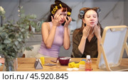 Two carefree adult besties sitting with colorful hair rollers on heads and applying nourishing facial mask to faces, enjoying self care and pampering moment at home together. Стоковое видео, видеограф Яков Филимонов / Фотобанк Лори