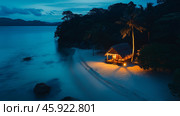 Romantic beachfront cottage illuminated at night surrounded by calm waters and palm trees in a tropical paradise. Стоковое AI-изображение, AI-художник Михаил Коханчиков / Фотобанк Лори