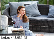 A woman is relaxing on the veranda of a country house and reading a book over a glass of red alcoholic drink or wine. The concept of relaxation and luxury wine. Стоковое фото, фотограф Photojuli / Фотобанк Лори