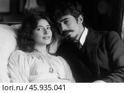 The black and white image shows a young couple, probably in love. The woman has long wavy hair, a soft gaze and a slight smile on her face. She is wearing a dress with a lace collar in the style of the early 20th century. The man next to her wears a suit with a tie and has a neat beard and moustache. Their pose emphasizes their closeness and trust in each other. The light is soft, creating a feeling of coziness and romance. Стоковое AI-изображение, AI-художник Анна Кривицкая / Фотобанк Лори
