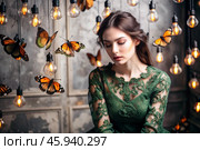 Portrait of a beautiful girl in a green dress among many suspended light bulbs and butterflies fluttering around her. Daydreaming. A magical atmosphere. Стоковое AI-изображение, AI-художник Photojuli / Фотобанк Лори