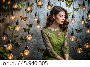 Portrait of a beautiful girl in a green dress among many suspended light bulbs and butterflies fluttering around her. Daydreaming. A magical atmosphere. Стоковое AI-изображение, AI-художник Photojuli / Фотобанк Лори