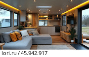 A modern interior of a mobile home or small living space, decorated in a minimalist and cozy style. The central place is occupied by a corner sofa with gray upholstery, decorated with decorative pillows of orange and neutral shades. Next to the sofa is a small wooden table. Opposite the sofa is a built-in TV and a storage cabinet with open shelves.The kitchen area is equipped with light cabinets with wooden accents, a built-in hob, a hood and built-in appliances. Decorative lighting of a warm yellow shade emphasizes the stylish design of the room and creates a cozy atmosphere. Large windows are installed on the walls, letting in natural light and opening up a view of nature. Стоковое AI-изображение, AI-художник Анна Кривицкая / Фотобанк Лори