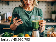 An elderly woman in the kitchen is looking at healthy food recipes on her phone and preparing a vegetable smoothie made from broccoli in a blender. Healthy detox diet drink. Стоковое AI-изображение, AI-художник Photojuli / Фотобанк Лори