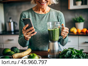 An elderly woman in the kitchen is looking at healthy food recipes on her phone and preparing a vegetable smoothie made from broccoli in a blender. Healthy detox diet drink. Стоковое AI-изображение, AI-художник Photojuli / Фотобанк Лори