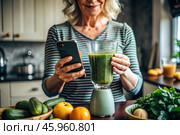 An elderly woman in the kitchen is looking at healthy food recipes on her phone and preparing a vegetable smoothie made from broccoli in a blender. Healthy detox diet drink. Стоковое AI-изображение, AI-художник Photojuli / Фотобанк Лори