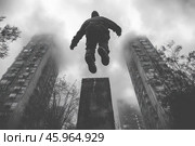 In black and white and conveys the dynamics of a person doing parkour. The central element of the scene is the figure of a man in a jump, captured in the air at the moment when he jumps from one surface to another. The man is dressed in dark clothes with a hood, which emphasizes his concentration and anonymity. In the background are two multi-story buildings, partially hidden by thick fog, creating an atmosphere of gloom and mystery. At the bottom of the composition, a concrete platform is visible, from which the jump begins. Trees with bare branches add a sense of severity and realism to the urban landscape. The photograph conveys the spirit of freedom, overcoming obstacles and determination. Стоковое AI-изображение, AI-художник Анна Кривицкая / Фотобанк Лори