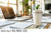 White paper coffee cup stands prominently on a desk filled with documents and a laptop, bathed in warm morning light, inviting focus and creativity for the day's tasks. Copy space. Стоковое AI-изображение, AI-художник Юлия Ильина / Фотобанк Лори