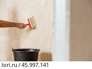 A hand with a wide brush and a bucket next to the wall being repaired during the application of primer or glue before gluing wallpaper. Wall treatment with a primer. Стоковое фото, фотограф Photojuli / Фотобанк Лори