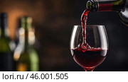 Red wine being poured into a glass with blurred wine bottles in the background during an evening gathering. Стоковое AI-изображение, AI-художник Сергей Толмачёв / Фотобанк Лори