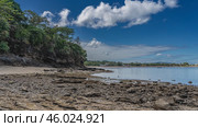 Low tide in the ocean. Boulders are scattered on the exposed seabed. Green vegetation on a coastal cliff. Boats and buildings on the shore are visible in the distance. Blue sky, clouds. Madagascar. Стоковое фото, фотограф Вера Ивановна Кубарева / Фотобанк Лори