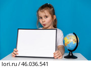 Happy little preschool girl sitting at table with empty white mock up paper board and a globe on a light blue background, copy space. Preschool education, traveling with children. High quality photo. Стоковое фото, фотограф Ирина Михайличенко / Фотобанк Лори