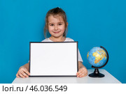 Happy little preschool girl sitting at table with empty white mock up paper board and a globe on a light blue background, copy space. Preschool education, traveling with children. High quality photo. Стоковое фото, фотограф Ирина Михайличенко / Фотобанк Лори