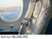 Airplane seat belt fastened over empty seat next to window with view of clouds at sunset during flight. Стоковое AI-изображение, AI-художник Иван Рябоконь / Фотобанк Лори