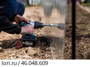 A man holds a battery-powered screwdriver and assembles a new greenhouse made of polyethylene, polycarbonate. Macro photography. Side view. Preparing for the spring garden season. Стоковое фото, фотограф Photojuli / Фотобанк Лори