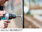 A man holds a battery-powered screwdriver and assembles a new greenhouse made of polyethylene, polycarbonate. Macro photography. Side view. Preparing for the spring garden season. Стоковое фото, фотограф Photojuli / Фотобанк Лори