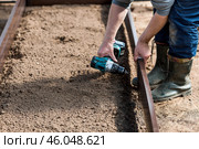 A man holds a battery-powered screwdriver and collects new beds from WPC into a greenhouse made of polyethylene, polycarbonate. Macro photography. Side view. Preparing for the spring garden season. Стоковое фото, фотограф Photojuli / Фотобанк Лори