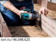 A man holds a battery-powered screwdriver and collects new beds from WPC into a greenhouse made of polyethylene, polycarbonate. Macro photography. Side view. Preparing for the spring garden season. Стоковое фото, фотограф Photojuli / Фотобанк Лори