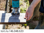 A man holds a battery-powered electric saw and builds new beds in a greenhouse made of wood-polymer composite. Macro photography. Side view. Preparing for the spring garden season. Стоковое фото, фотограф Photojuli / Фотобанк Лори