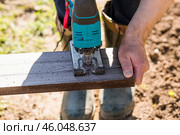 A man holds a battery-powered electric saw and builds new beds in a greenhouse made of wood-polymer composite. Macro photography. Side view. Preparing for the spring garden season. Стоковое фото, фотограф Photojuli / Фотобанк Лори