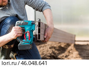 A man holds a battery-powered electric saw and builds new beds in a greenhouse made of wood-polymer composite. Macro photography. Side view. Preparing for the spring garden season. Стоковое фото, фотограф Photojuli / Фотобанк Лори