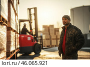 African American warehouse worker in his 30s, standing by stacked pallets, with forklift operating in background during sunrise. Стоковое AI-изображение, AI-художник Владимир Григорьев / Фотобанк Лори